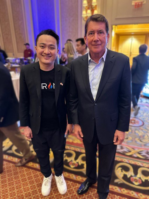 Two men pose for a photo at what appears to be a conference or networking event in an upscale venue with ornate carpeting. The younger man on the left wears a black blazer over a t-shirt displaying the "RON" ticker/logo, suggesting a connection to the Ronin or related crypto project, while the older man on the right wears a formal dark suit. The image visually complements the post's theme of crypto conviction, likely showing a notable industry figure encounter that reinforces the author's commitment to staying concentrated in crypto rather than diversifying into traditional assets.