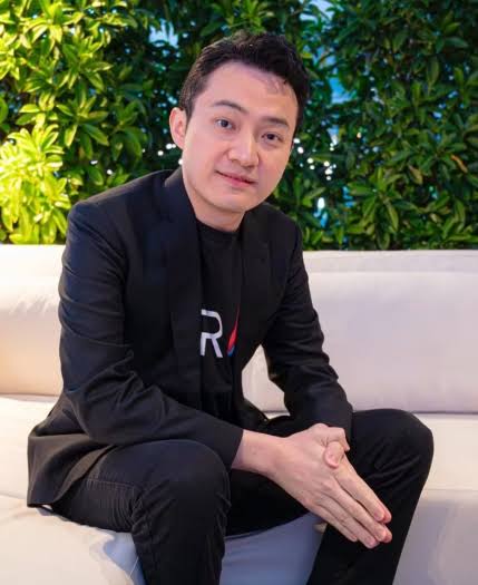 The image shows Justin Sun (Tron founder) seated on a white couch in a black blazer over a black t-shirt featuring the red "TRX" Tron logo, with his hands clasped in front of him against a backdrop of green foliage, accompanying a post highlighting his evolving career trajectory and expanding ventures by 2025.
