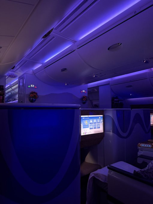 A luxurious business-class cabin bathed in moody purple ambient lighting, featuring a seatback entertainment screen displaying "15" alongside other flight options. The premium setting visually mirrors the poster's celebratory mood of affording business-class travel thanks to Bitcoin's recent price surge.