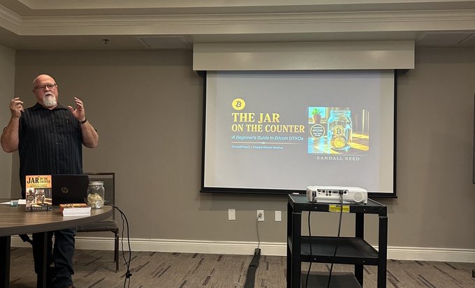 A man with a white beard and glasses stands presenting in a room next to a projection screen displaying 'THE JAR ON THE COUNTER: A Beginner's Guide to Bitcoin UTXO' by Randall Reed.