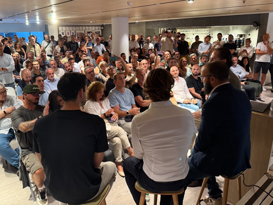 A photo of a large audience attending an indoor event or talk, with panelists seated at the front and attendees listening attentively.