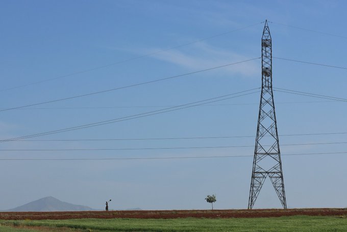 A lone figure stands on a vast grassy plain beside a small tree, dwarfed by a towering electrical transmission pylon and set against distant mountains under a bright blue sky—a serene composition reflecting the harmony between humanity and nature featured in the Tezos photography collection.