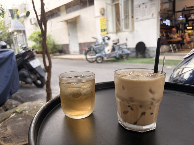 Two beverages—a glass of light tea or beer and an iced coffee—rest on a tray at a sidewalk café, with scooters and a street scene visible in the background. The caption evokes a relaxing pause in the day, fitting the post's cheerful message wishing friends a happy #DogeDay Tuesday evening.
