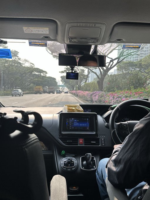 A view from the passenger seat of a moving car, showing the driver, dashboard, and a road ahead with other vehicles.