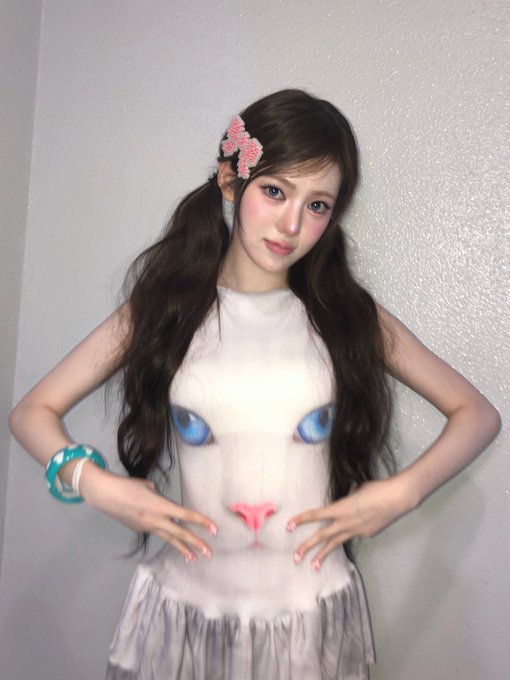 A young woman with long dark wavy hair wears a white tank top printed with a cat face design and a pink bow hair clip, posing against a light wall.