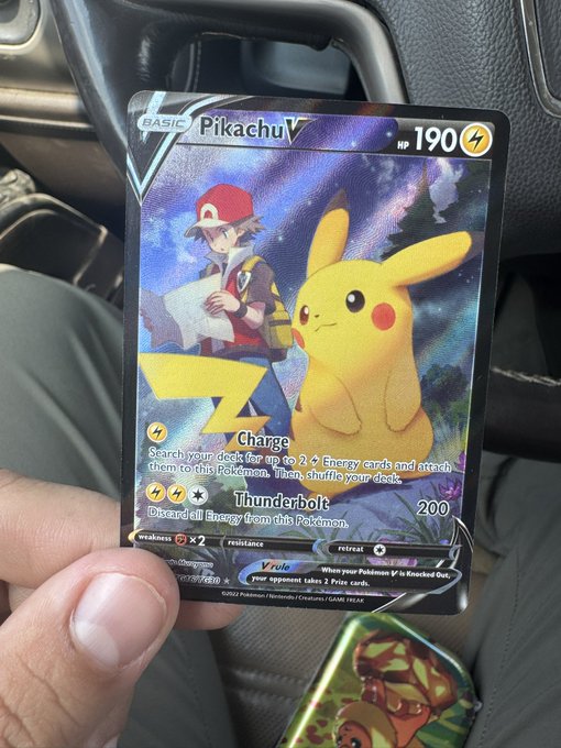 A hand holding a holographic Pikachu V Pokémon card showing the 'Charge' and 'Thunderbolt' attacks, with Pikachu and Ash Ketchum artwork, displayed inside a car.