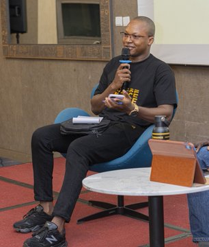 A man wearing glasses and a black t-shirt with yellow text sits in a blue chair holding a microphone and a phone, speaking at what appears to be a panel or event.