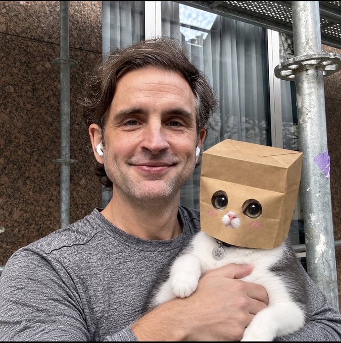 A man with AirPods wearing a gray shirt smiling while holding a white and gray cat wearing a brown paper bag with cutout eyes and drawn-on cat features over its head.