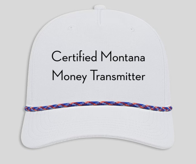 A white baseball cap with red and blue patterned rope trim displays the text "Certified Montana Money Transmitter," suggesting it's a novelty item that could be sold in the Colossus merch shop.