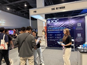 The image shows a group of people gathered around a booth at an event. The booth belongs to XDC Network, which is hosting a conference for blockchain enthusiasts. A woman stands behind the booth, talking to attendees while they look at her presentation materials. There are several people in the scene, including some standing closer to the booth and others further away.

The background of the image features a large banner that reads "XDC Network" along with the words "layer chain", indicating the focus of the event is on blockchain technology. The attendees appear engaged and interested in what the woman at the booth has to say, suggesting that this conference might be an important gathering for those who are passionate about blockchain and its applications.