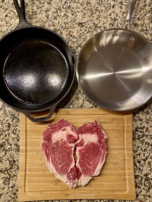 The image shows a cutting board with two pieces of raw meat, possibly steak or beef, placed on top of it. There are also two pans - one black and one silver - sitting next to the cutting board. The background appears to be a kitchen countertop.