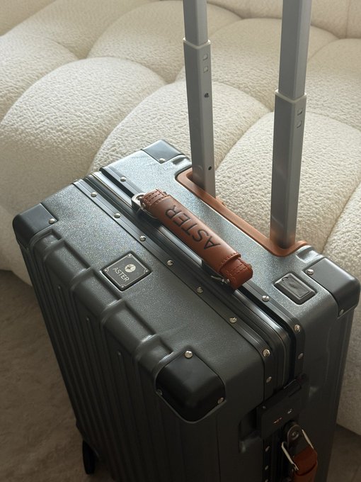 A black Aster aluminum-frame carry-on suitcase sitting on a textured beige surface next to a white cushion.