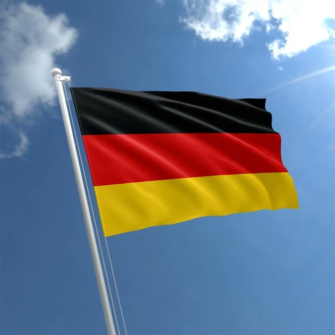 A photograph of the German national flag (black, red, and gold horizontal stripes) waving on a white flagpole against a bright blue sky with scattered white clouds.