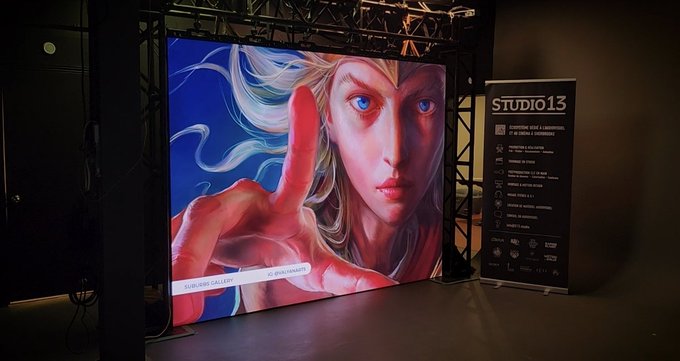 A large digital display screen at 'STUDIO 13' showcasing digital art of a blonde anime-style character with blue eyes pointing forward, with text reading 'SUBIRÁN GALERÍA' and '@subirangallery', alongside an information panel listing services like crypto art gallery, photography, and beauty salon.