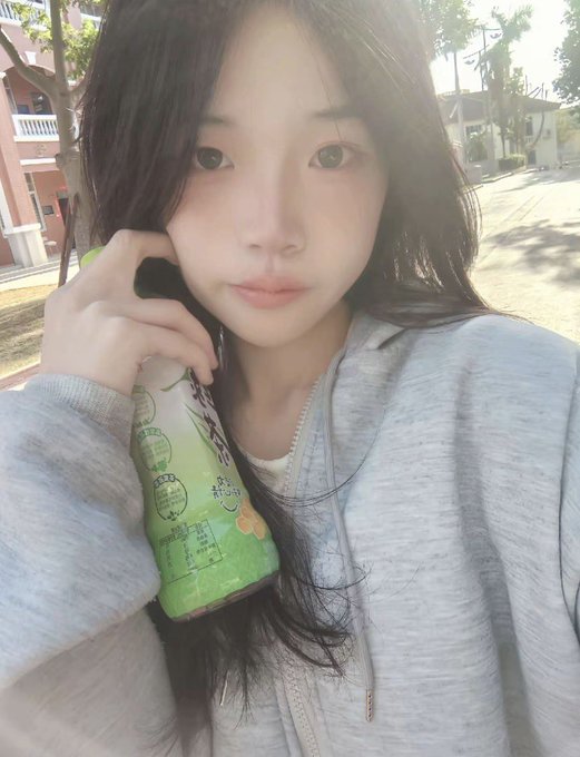 The image features a young woman with black hair wearing a gray sweatshirt holding a green bottle of soda. She has her eyes closed while posing for the photo. The background includes trees and buildings, suggesting that she might be outdoors or near an urban area. The caption reads "Today, as usual, sending GM to my friends! A simple good morning hides a whole night of sleeplessness To others, it' s just an ordinary morning greeting Only I know I endured the long, endless night Staring at the market fluctuations the entire time, staying alert to every".