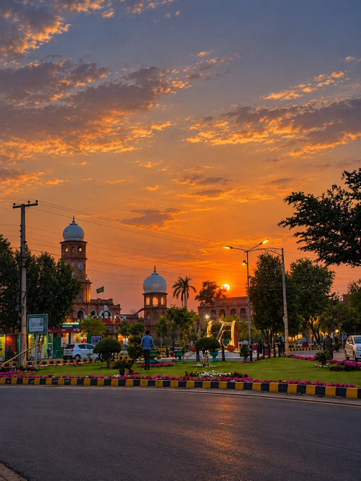 A vibrant sunset scene over a city street, with the sun setting behind a mosque-like building featuring blue domes, casting orange and pink hues across the cloudy sky.