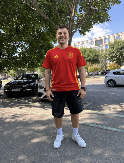 A young man in a red Spain national team soccer jersey, black shorts, and white sneakers standing outdoors near parked cars.