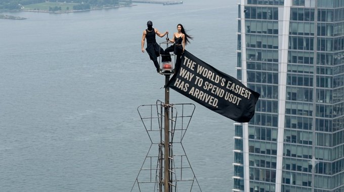 Two people sitting atop a tall pole with a black banner reading 'THE WORLD'S EASIEST WAY TO SPEND USDC HAS ARRIVED,' overlooking a body of water with a high-rise building in the background.