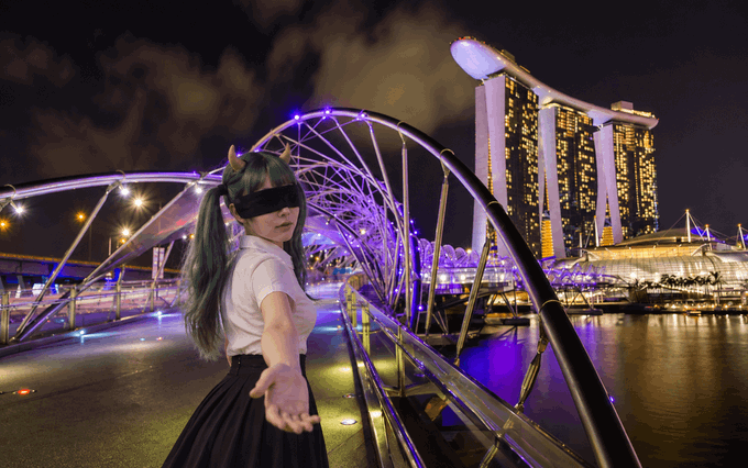 A person with teal pigtails and a black blindfold reaches out on an illuminated bridge at night, with the distinctive Marina Bay Sands hotel and a Ferris wheel visible in the background.