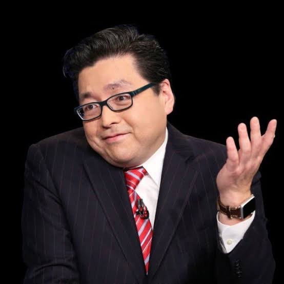 A man in a dark pinstripe suit, white shirt, and red-striped tie wears glasses and gestures with his raised left hand against a black background, smiling toward the camera.