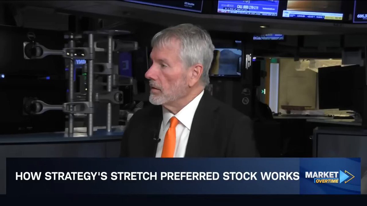 The image shows a man wearing an orange tie standing in front of a computer screen with a news article about strategy's preferred stock works posted above him. The text on the screen reads "How Strategy's Stretch Preference Works".