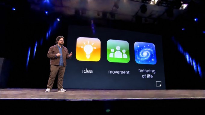 A bearded man stands on stage presenting in front of a large screen displaying three colorful app-style icons labeled "idea," "movement," and "meaning of life," echoing the post's reference to e/acc as an idea, a movement, and the meaning of life.
