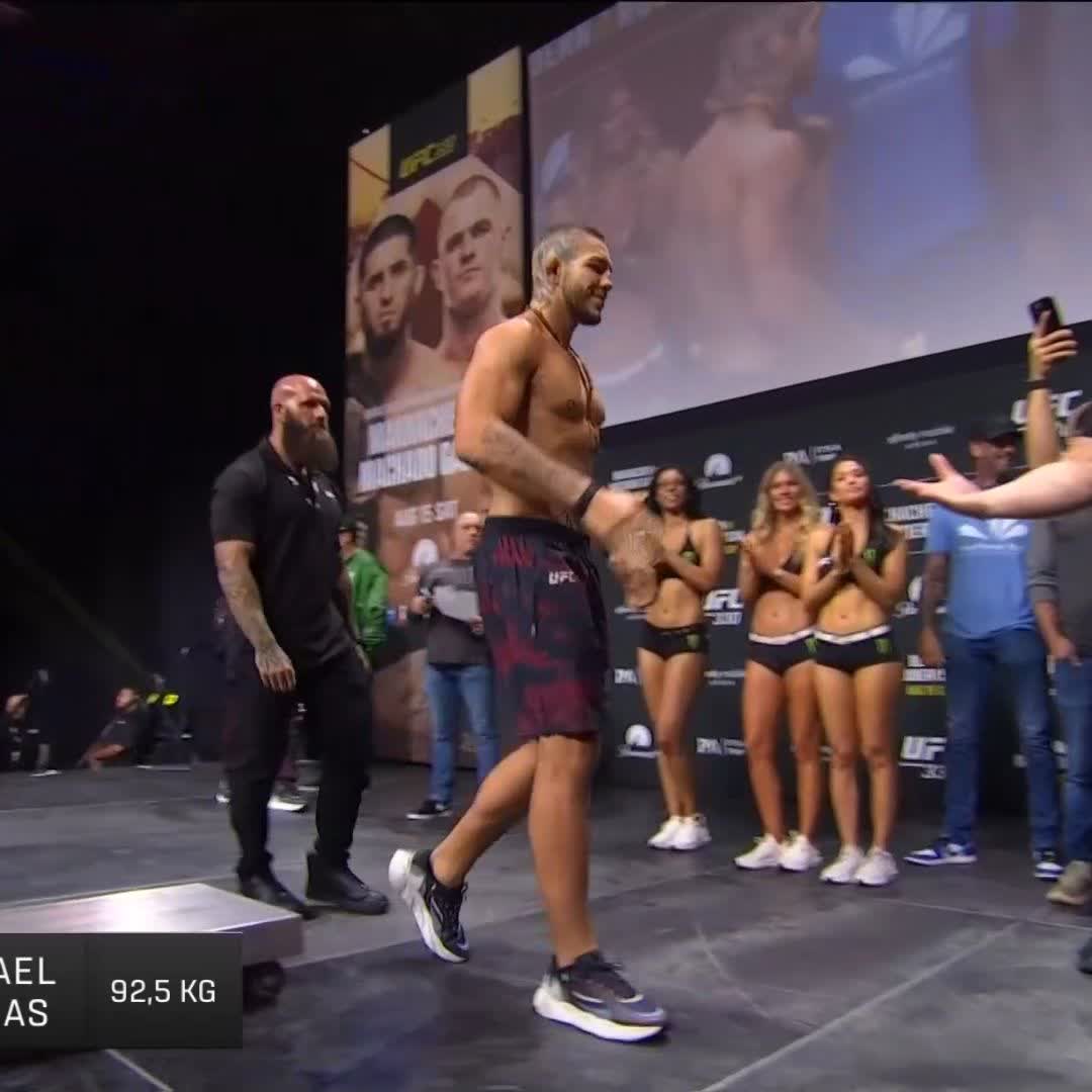A shirtless UFC fighter walks onto the stage during the weigh-in, with "92,5 KG" displayed on the screen as part of the UFC 330 event, accompanied by ring girls and security personnel. The image captures the pre-fight weigh-in moment featuring the Brazilian matchup between the fighters mentioned in the post.