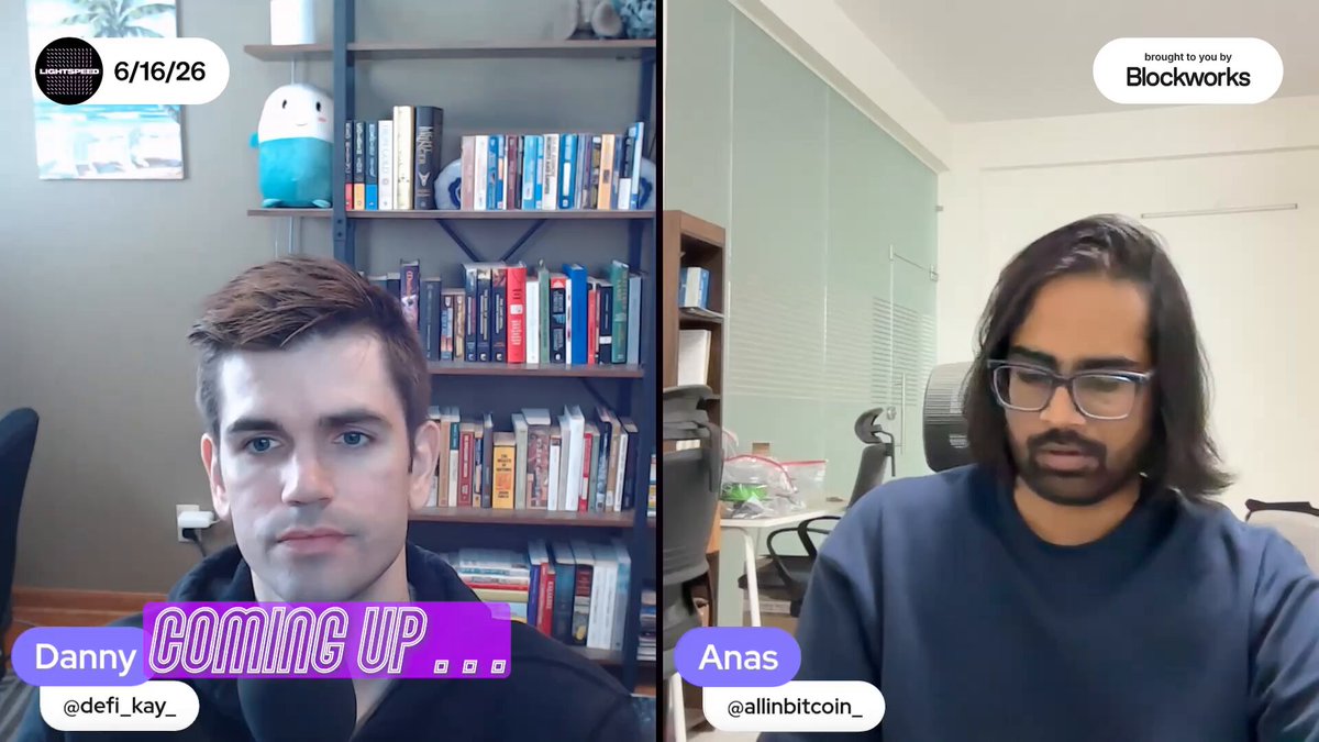 The image shows two men sitting at a desk, having a video call with each other. They are both wearing glasses and appear to be engaged in conversation. The background of the image reveals that they are in an office setting, as there are several books scattered around them on shelves or tables.