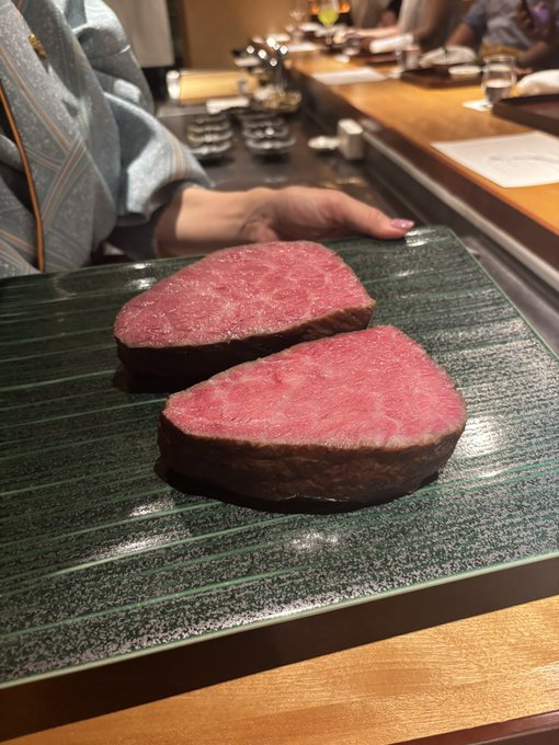 Two thick cuts of rare, seared beef steak are presented on a green textured plate at a teppanyaki restaurant, with a chef visible in the background.