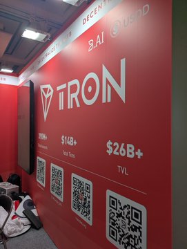 The image shows the TRON booth display featuring the company's logo alongside key statistics: 220M+ users, $14B+ in Total Value, and $26B+ TVL (Total Value Locked), with QR codes visible below for downloading the app. This relates to the post as the visitor stopped by the TRON booth at what appears to be a conference or convention, highlighting their engagement with the TRON ecosystem led by Justin Sun.