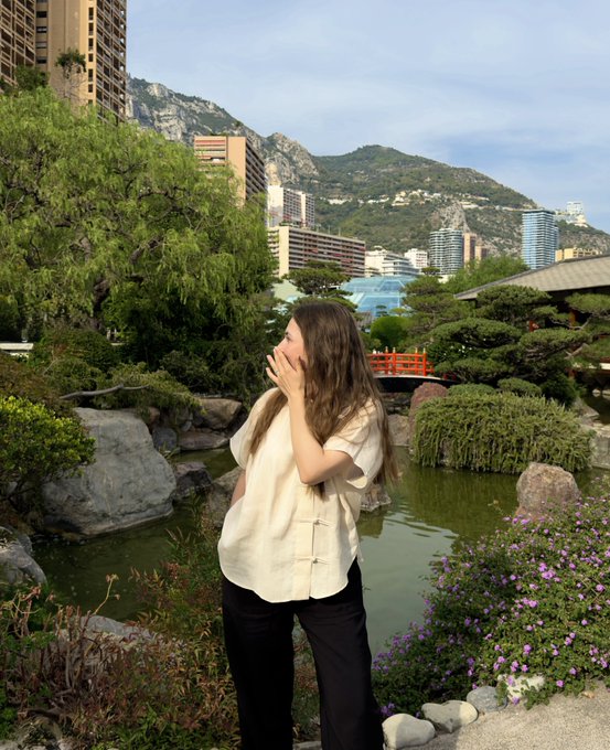 A woman with long brown hair wearing a cream-colored blouse and black pants stands in a Japanese-style garden with a pond, rocks, and a small red bridge, with modern buildings and mountains visible in the background.