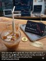 A dimly lit scene of a person drinking an iced beverage at a table with a tablet displaying a stock chart and a plate of pancakes in the background.