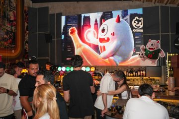 A lively crowd gathers at a bar featuring a large illuminated screen displaying a cartoon character with red horns and a tiger mascot logo marked "GOMU," set against the backdrop of an evening event at the Istanbul Şişli Bomonti Hotel. The scene captures the festive atmosphere of the TRON Global Friends VIP World Cup Final Event, with attendees socializing beneath colorful lighting and the venue's vibrant decor.
