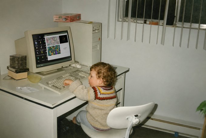 A vintage photograph of a young child with curly hair sitting at a desk using an old Macintosh computer displaying colorful graphics.