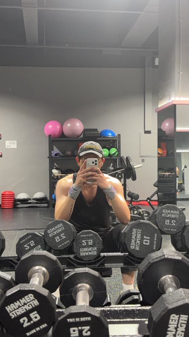 A man in a tank top and cap takes a mirror selfie in a gym, surrounded by Hammer Strength dumbbells marked "2.5" and "10," with his phone obscuring his face. The post's request to discuss crypto/finance data parallels the visual "lifting heavy" theme—both being relatable struggles of handling weight, whether iron or portfolio.