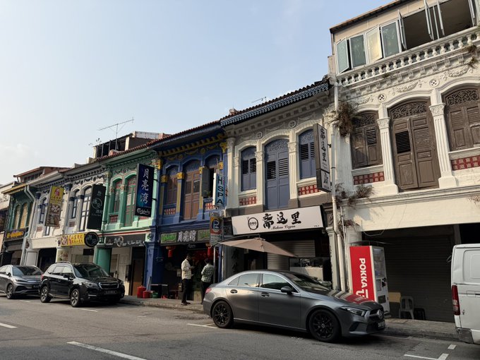 A row of colorful shophouses lines a street in Singapore, featuring storefronts with Chinese signage including "月泉湾" and "东马里," alongside parked cars and a few pedestrians on the sidewalk.