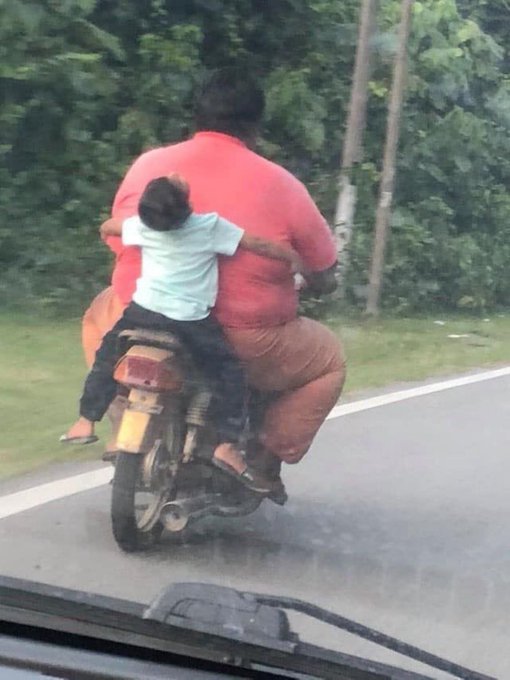 A candid photo taken from inside a car showing a large woman in a salmon shirt riding a motorcycle with a small child in a white shirt clinging to her back on a tree-lined road.