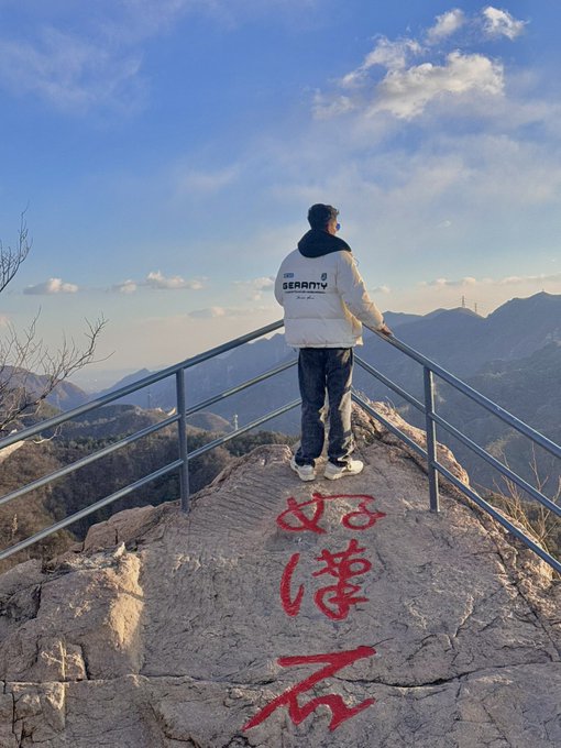 A person wearing a white 'SECURITY' jacket stands at a mountain viewpoint railing looking out over the landscape, with red Chinese characters painted on the rock below.