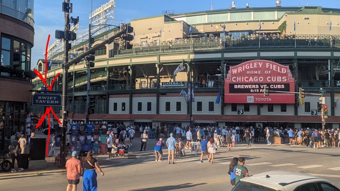 An exterior view of Wrigley Field showing the marquee displaying 'Home of Chicago Cubs, Brewers vs Cubs, 6:15 PM, Toyota' with red arrows pointing to a 'Swift Tavern' sign.