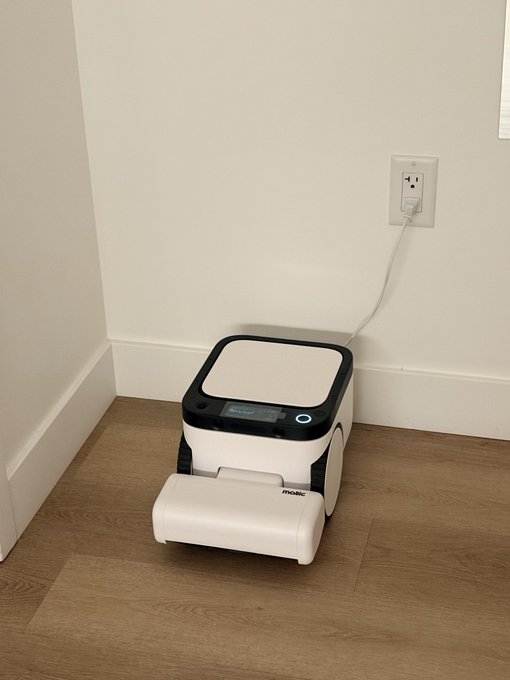 A modern home scene showing a white and black Matic robot vacuum docked at its charging station, plugged into a wall outlet on a wooden floor in the corner of a room.