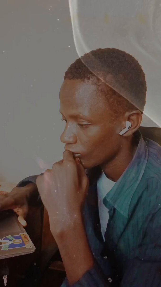 The image features a young man with short hair wearing a blue shirt, sitting at a desk or table while using his cell phone. He has an earbud in one ear and appears to be engaged in some activity on his phone. The background is blurred out, drawing attention to the main subject of the photo.
