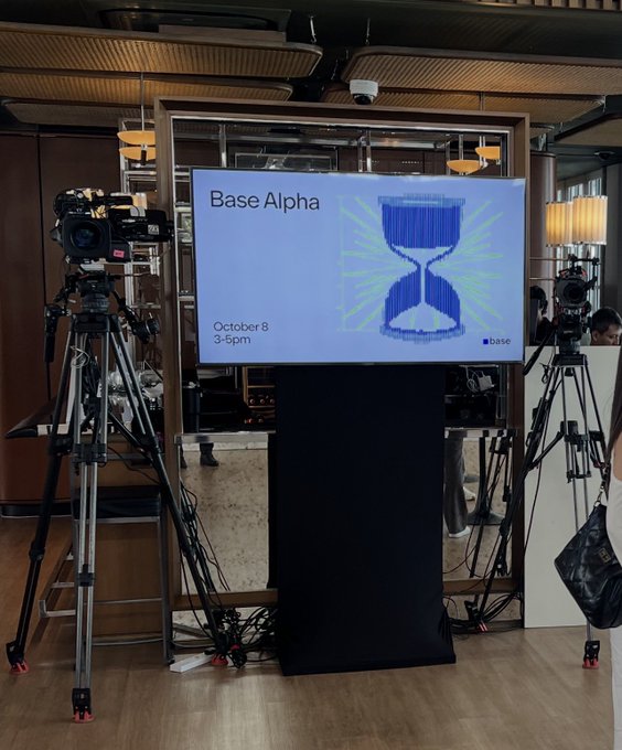 A television display showing "Base Alpha October 8 3-5pm" with an hourglass graphic, flanked by two professional video cameras on tripods set up in what appears to be a studio or event space.