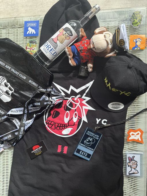 A flat lay of Bored Ape Yacht Club merchandise including a black hoodie with a red skull logo, a cap, lanyards, stickers, a bottle of BORED Vodka, and a small monkey plush toy.