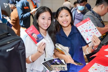 Two young women smile while holding promotional booklets featuring cartoon illustrations, with the red booklet in front appearing to display "MyBook." The Tron logo is visible in the top left corner, and the "See you next time" caption suggests this is a farewell moment at a recent event or gathering.