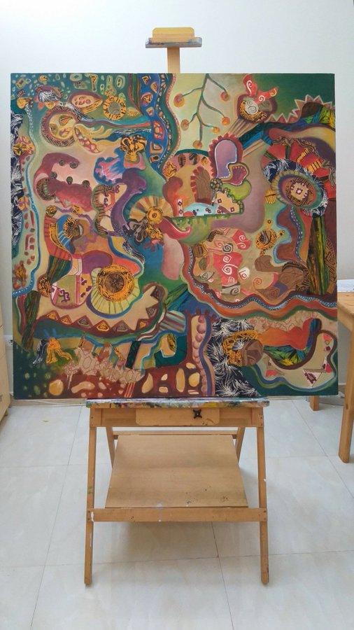 A vibrant, colorful abstract painting featuring organic, flowing shapes and figures in earthy and bright tones sits on a wooden easel in a well-lit room with white tile floors.