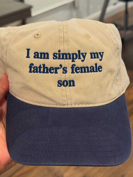 A beige and navy blue baseball cap held in a hand, with embroidered text reading "I am simply my father's female son".