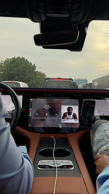 The image shows the interior of a vehicle, viewed from the backseat, with passengers enjoying music on the central touchscreen display, which shows what appears to be an artist named "Jorja W" playing a song titled "Waiting." A phone mounted next to the screen displays a navigation app, while a black pickup truck is visible ahead through the windshield, capturing a casual carpool moment that fits the relaxed "g m guys" vibe of the post.