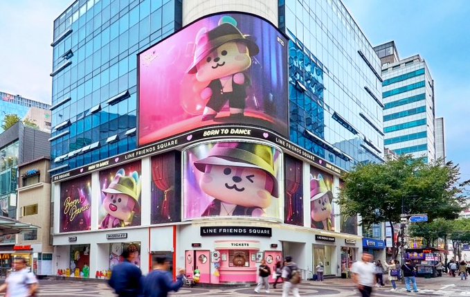 The exterior of LINE FRIENDS SQUARE featuring large LED screens displaying LINE Friends characters like Brown and Sally wearing colorful hats, with the slogan 'BORN TO DANCE'.