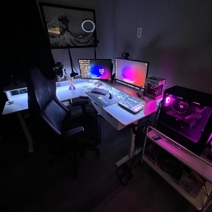 A dimly lit gaming/streaming setup featuring dual monitors, a keyboard, a microphone, and a PC case illuminated with pink and purple RGB lighting, accompanied by a gaming chair and a wave artwork on the wall.