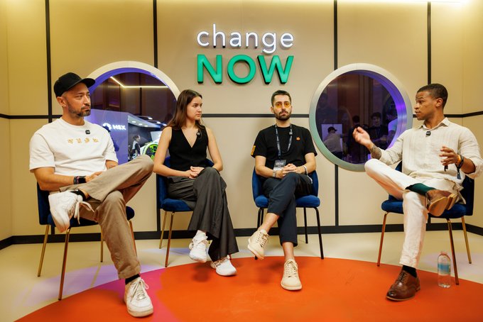 Four people sit on stools in front of a 'change NOW' sign for what appears to be a panel discussion at a corporate event.